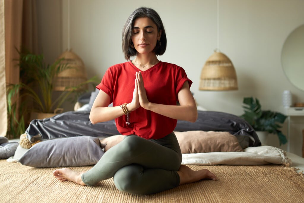 Fashionable young female with grayish hair sitting in gomukhasana or cow pose while practicing hatha yoga in the bedroom after awakening, keeping eyes closed, pressing hands together in namaste